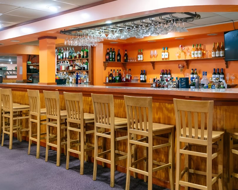 A long wooden bar with several empty wooden bar stools and shelves of liquor bottles.