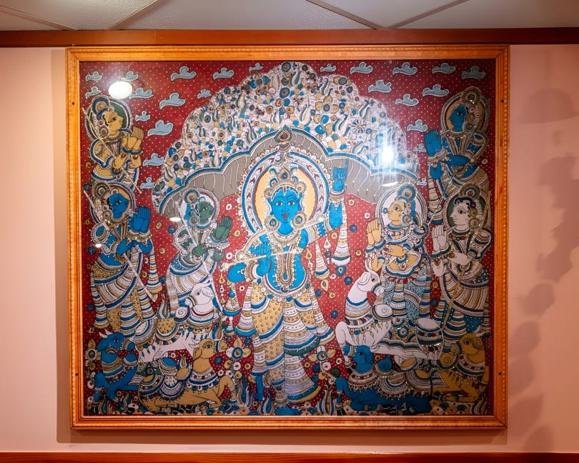 A framed, colorful Indian painting depicts a blue-skinned deity playing a flute, surrounded by figures and animals.