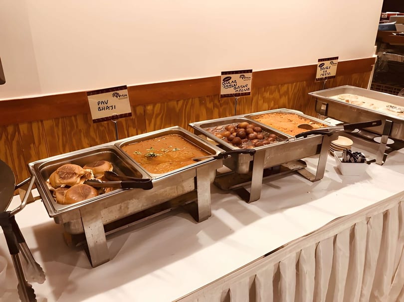 A buffet line with chafing dishes containing Pav Bhaji, Gulab Jamun, and other Indian dishes.