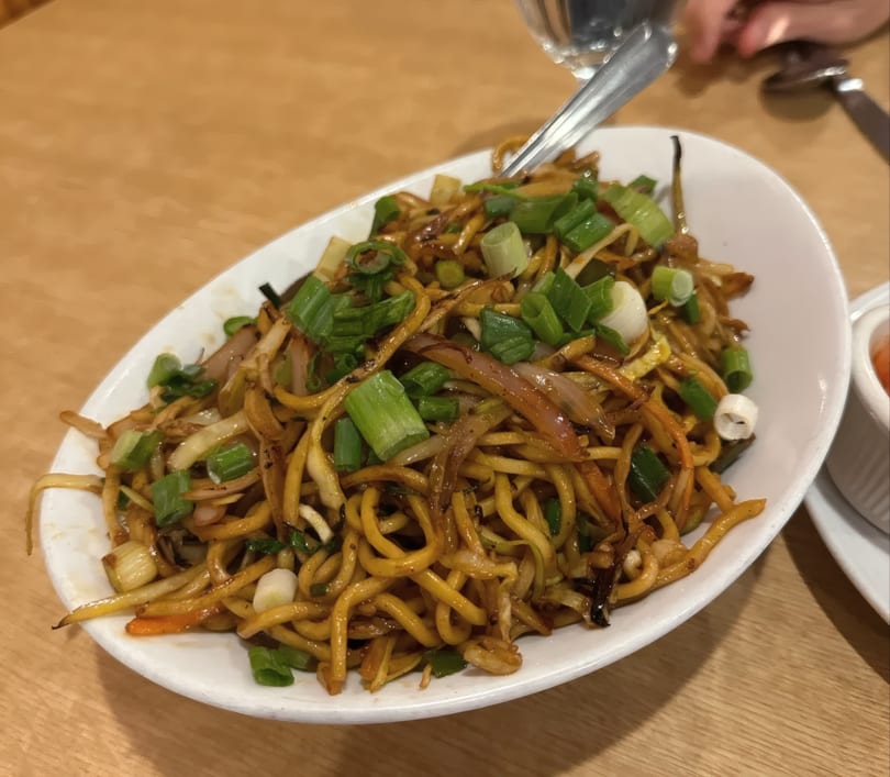 A white oval plate filled with stir-fried noodles, green onions, and vegetables on a wooden table.
