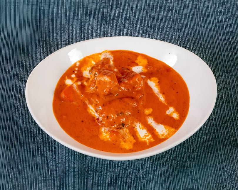 A white bowl filled with orange butter chicken, drizzled with white cream, on a dark blue placemat.