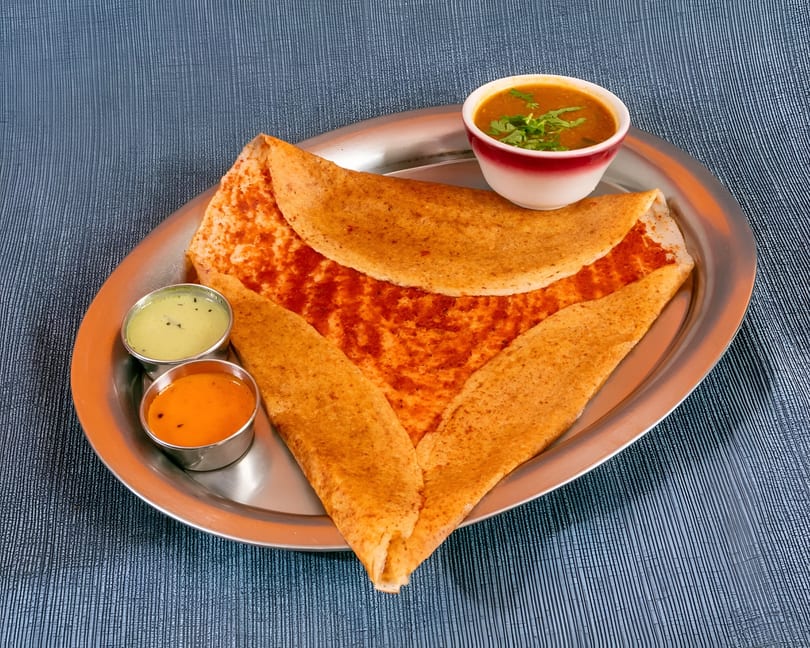 Masala dosa with sambar and two chutneys on a silver platter, against a blue textured background.