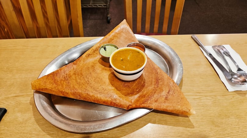 A golden-brown dosa, folded into a triangle, served on a metal plate with three small bowls of sauces.