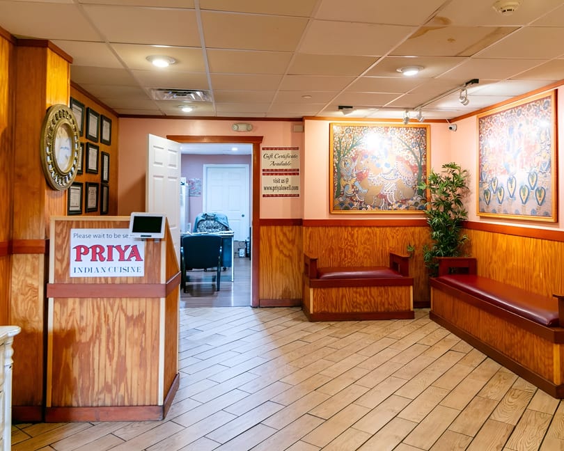 Priya Indian Cuisine restaurant waiting area with a host stand, wooden benches, and colorful Indian art.