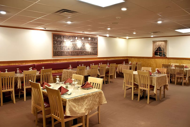 An empty restaurant dining room with many tables set with red napkins and light-colored tablecloths.