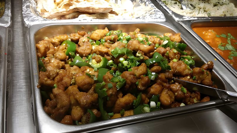 Buffet tray of chili chicken with green peppers and scallions, alongside naan and rice.
